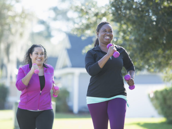 Two multi-ethnic women exercising outdoors, jogging on a sunny day in a residential neighborhood, handweights in their hands. The main focus is on the African American woman, laughing as her Hispanic friend tries to catch up with her.