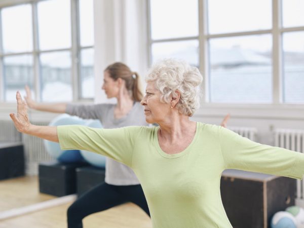 Two women doing stretching and yoga workout at gym. Female trainer in background with senior woman in front during physical training session