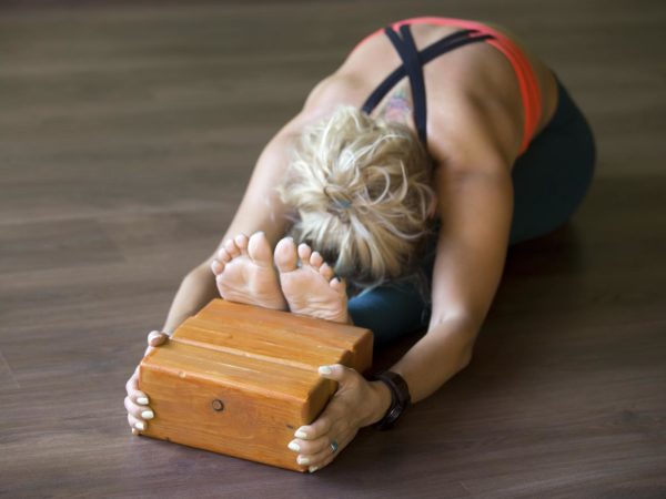 Sporty beautiful young blond woman in sportswear working out indoors, doing exercise for spine, shoulders, hamstrings, sitting in seated forward bend posture, focus on wooden bricks