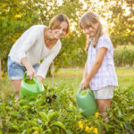 Mother and daughter watering vegetables in their garden. Trees and corn field visible in the background. Sunlit from the back.