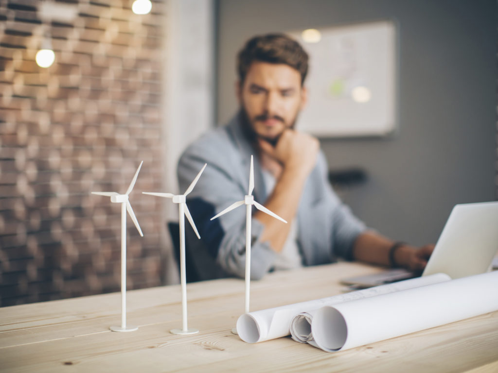 Young engineer at modern office space, sitting at the table and working on energy project. Focus on wind turbine models on the table.