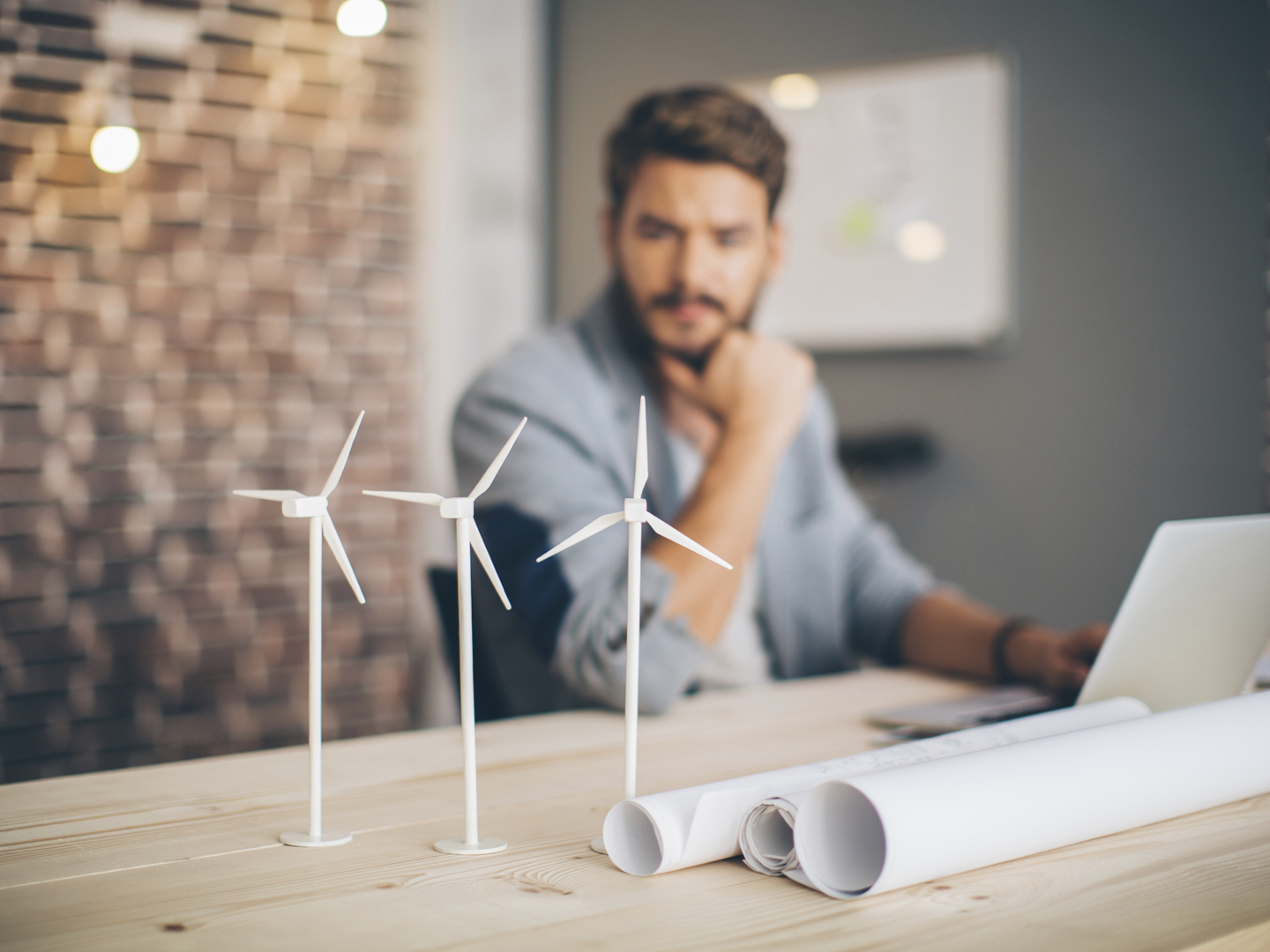 Young engineer at modern office space, sitting at the table and working on energy project. Focus on wind turbine models on the table.