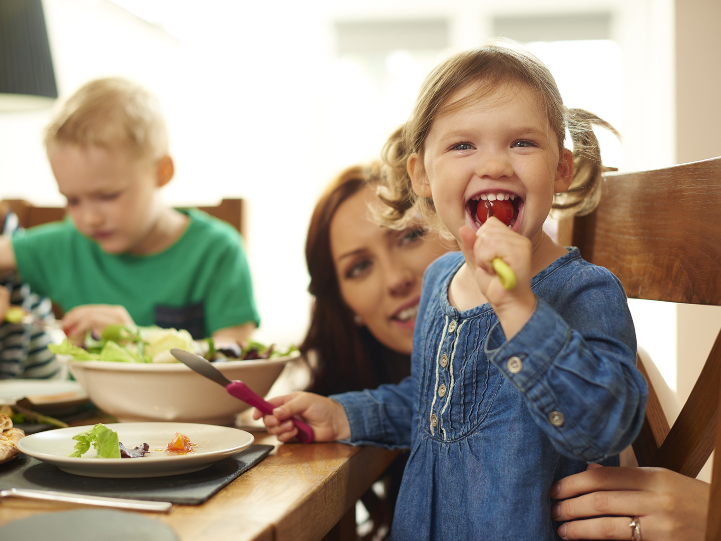 a young family of four sit at the dining table and eat pizza and salad.Mum helps her little girl with her meal , as she cheekily stuffs a whole tomato into her mouth .