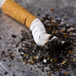 A white and tan colored cigarette butt with a filter extinguished inside a silver colored ashtray. There is ash all around the cigarette butt and the focus is on the end of the cigarette butt.