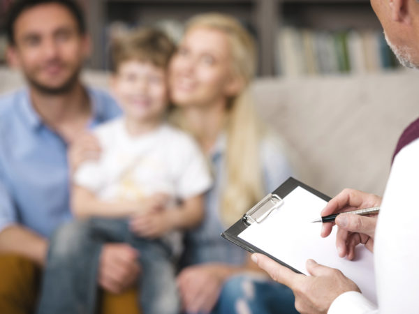 Young happy family of three talking with psychologist. Focus on psychologist. There are many books in psychologist office