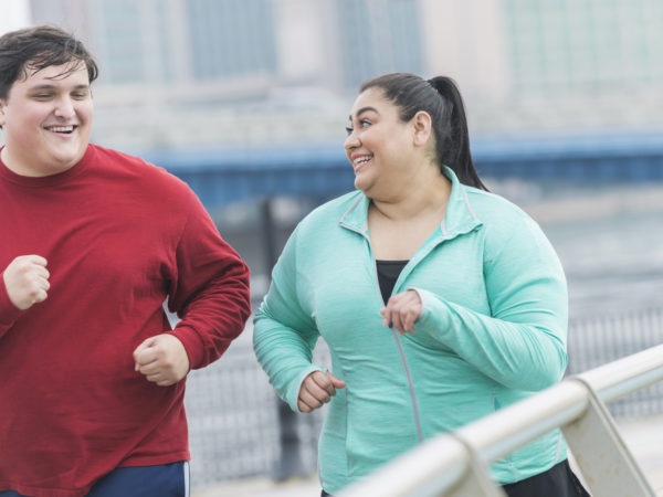 An overweight Hispanic woman and a young mixed race Hispanic and Caucasian man exercising together outdoors in an urban setting, running or jogging. They are smiling, looking at each other as they exercise.