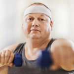 Mid adult overweight man exercising with dumbbells and looking at the camera.