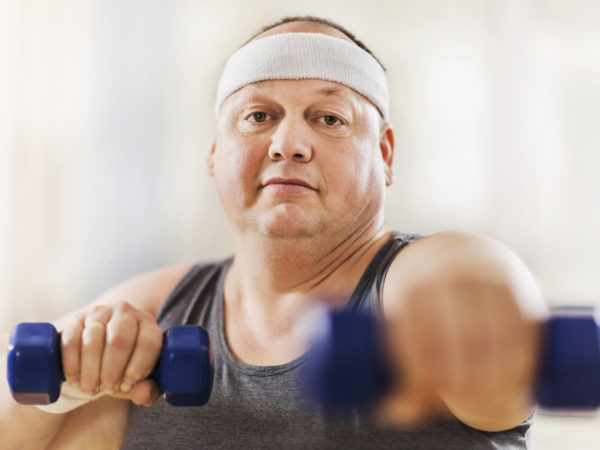 Mid adult overweight man exercising with dumbbells and looking at the camera.