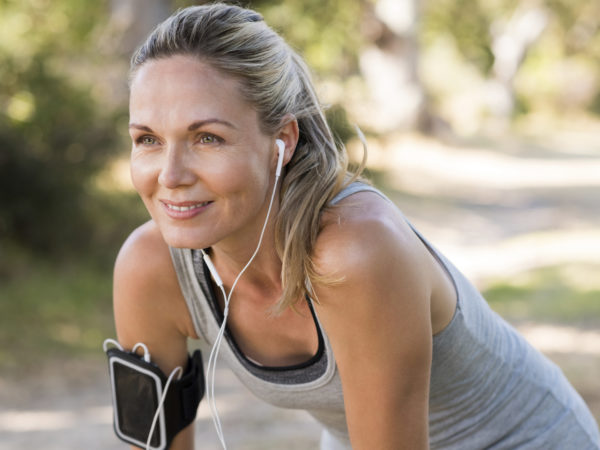 Portrait of athletic mature woman resting after jogging. Beautiful senior blonde woman running at the park on a sunny day. Female runner listening to music while jogging.