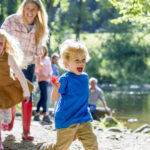 A family are sat at a lake on a nice summers day. The children are running ahead while parents look on.