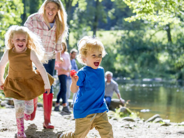 A family are sat at a lake on a nice summers day. The children are running ahead while parents look on.