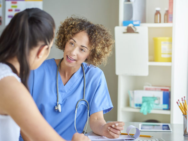 A female doctor sits at her desk and listens to a female patient .  She is wearing blue scrubs and stethoscope and is sitting at her desk . In the background shelving containing sharps box, gloves and medicine can be seen .