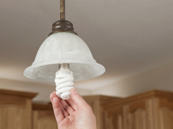 A male hand is installing an energy efficient compact fluorescent light bulb (CFL) into a ceiling hung pendant fixture. The background is kitchen cabinets with above cabinet lighting.