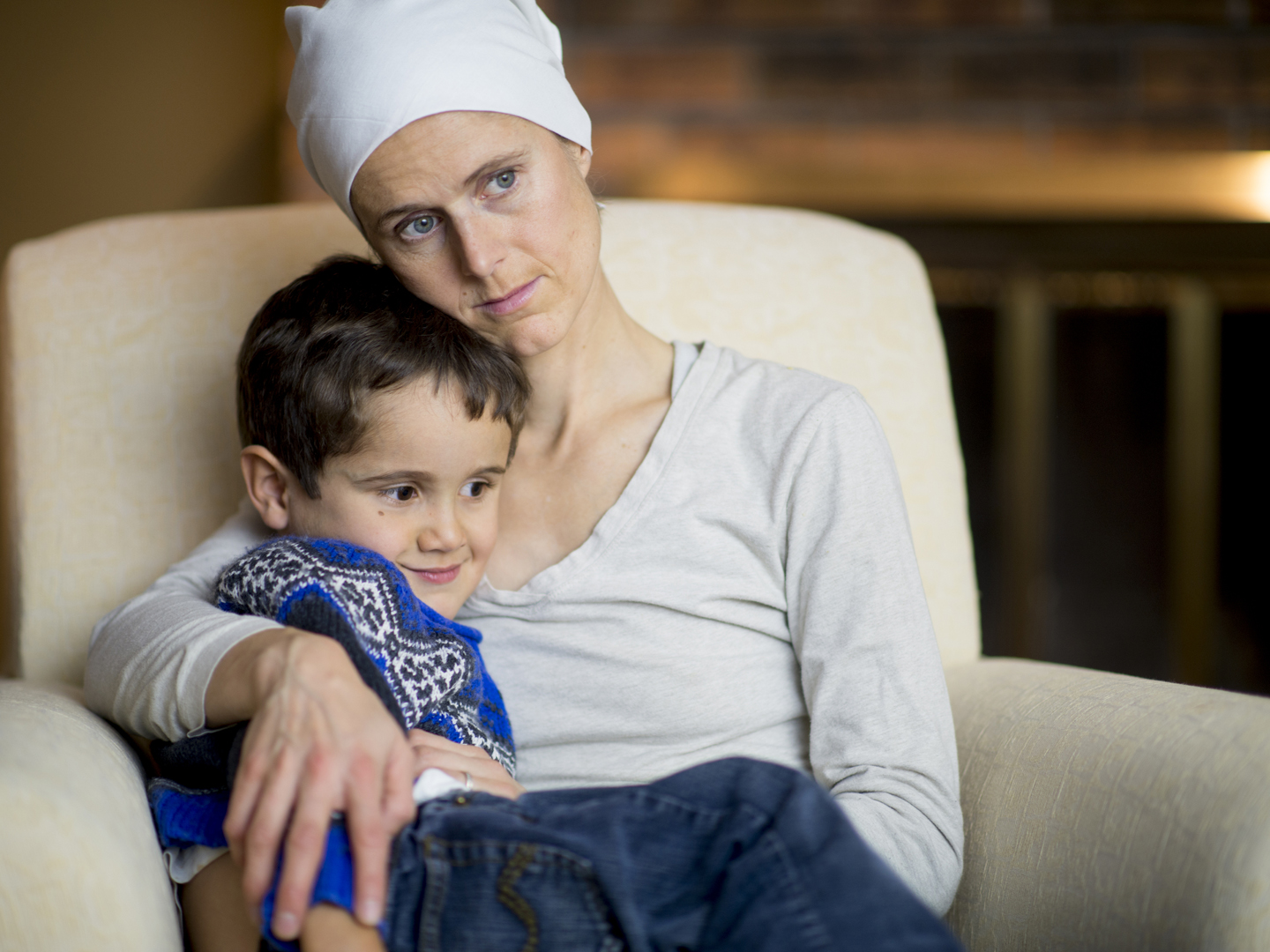 A mother with cancer has lost her hair from the chemotherapy and is wearing a bandana. She is sitting with her son on a chair in their home.