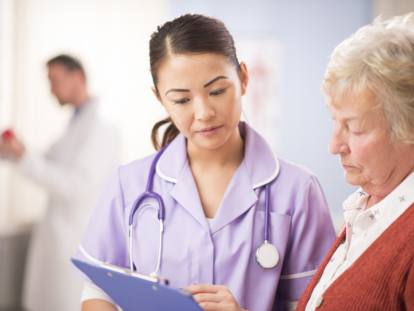 a young nurse stands and chats to a senior woman patient as she leaves the surgery. She is wearing a lilac nursing uniform and holding a clipboard . She is smiling to the senior woman and explaining her treatment plan. In the background a hospital interior can be seen. A male doctor is defocussed in the background.