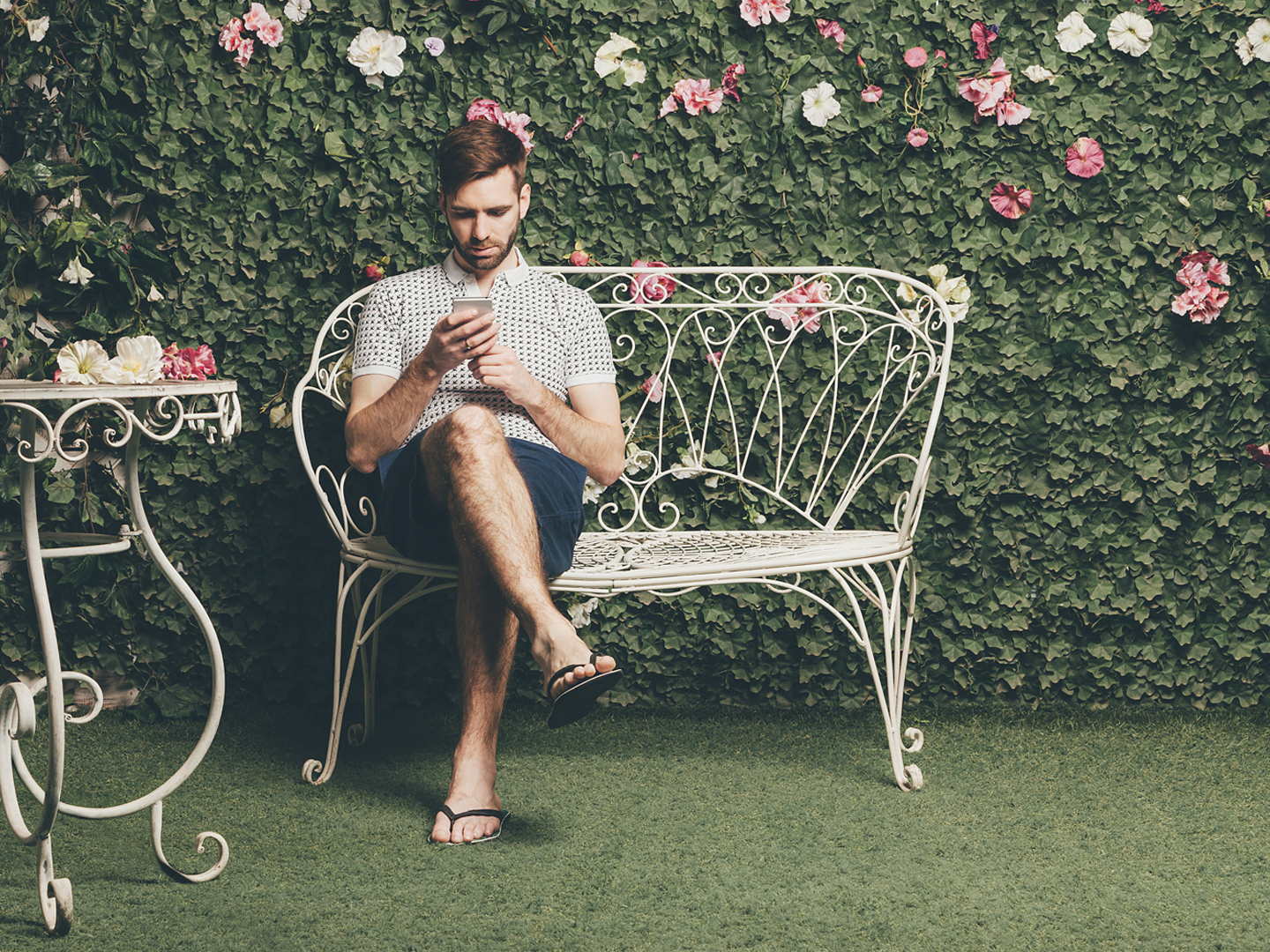 Handsome man sits on a vintage bench and typing text message, green wall of leaves and flowers on background.