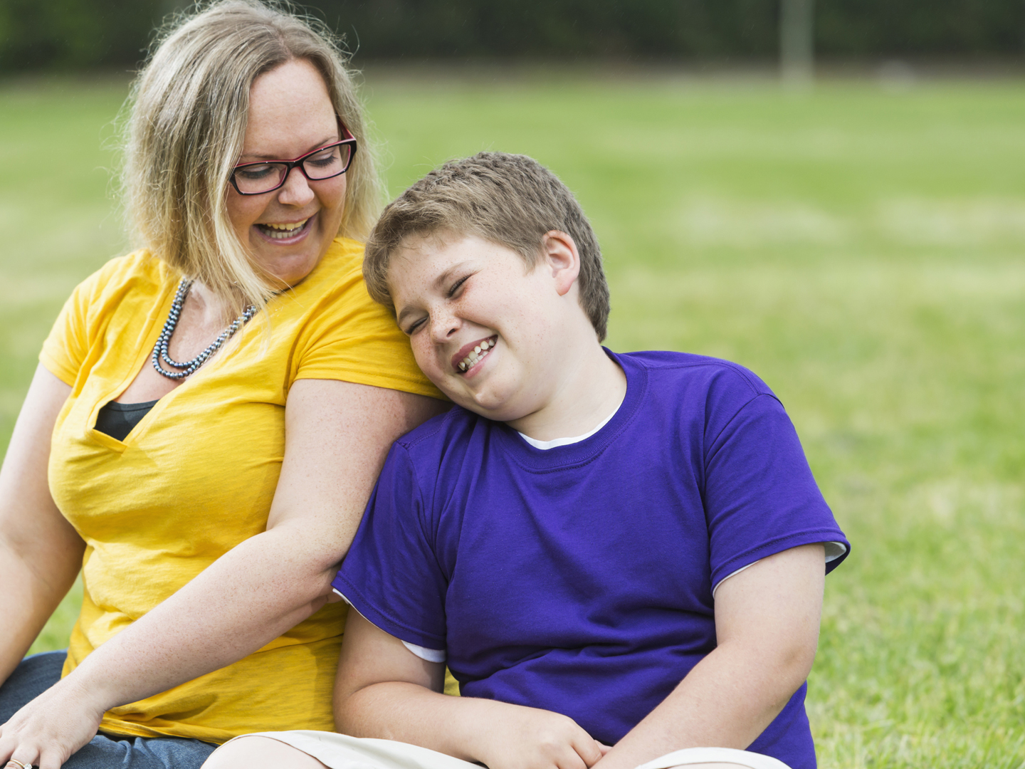 Mother and preteen son sitting together on the grass in the park, laughing. The boy is leaning his head against his mom's shoulder and she is looking down at him. They are wearing casual clothing, jeans, shorts, and t-shirts. They are ordinary people, heavyset, happy and confident. The woman is wearing eyeglasses.