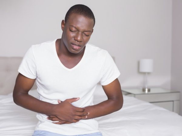 Wincing man sitting on the edge of his bed with a stomach cramp at home in bedroom