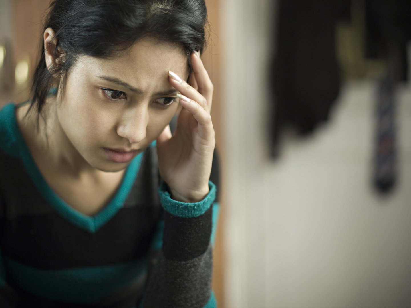 Indoor low key image of a serene, stressed young woman feeling headache and thinking by touching her head and looking down with blank expression. She is wearing a sweater. One person, waist up, horizontal composition with selective focus and copy space.