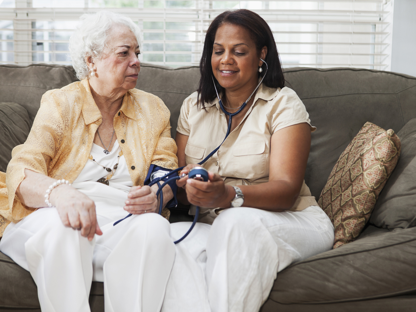 Senior Hispanic woman (70s) with caregiver (50s) at home, taking blood pressure.