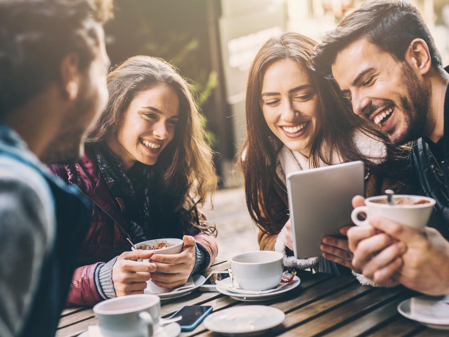 Laughing young people looking at tablet and laughing while sitting in cafe outdoors, holding cups.