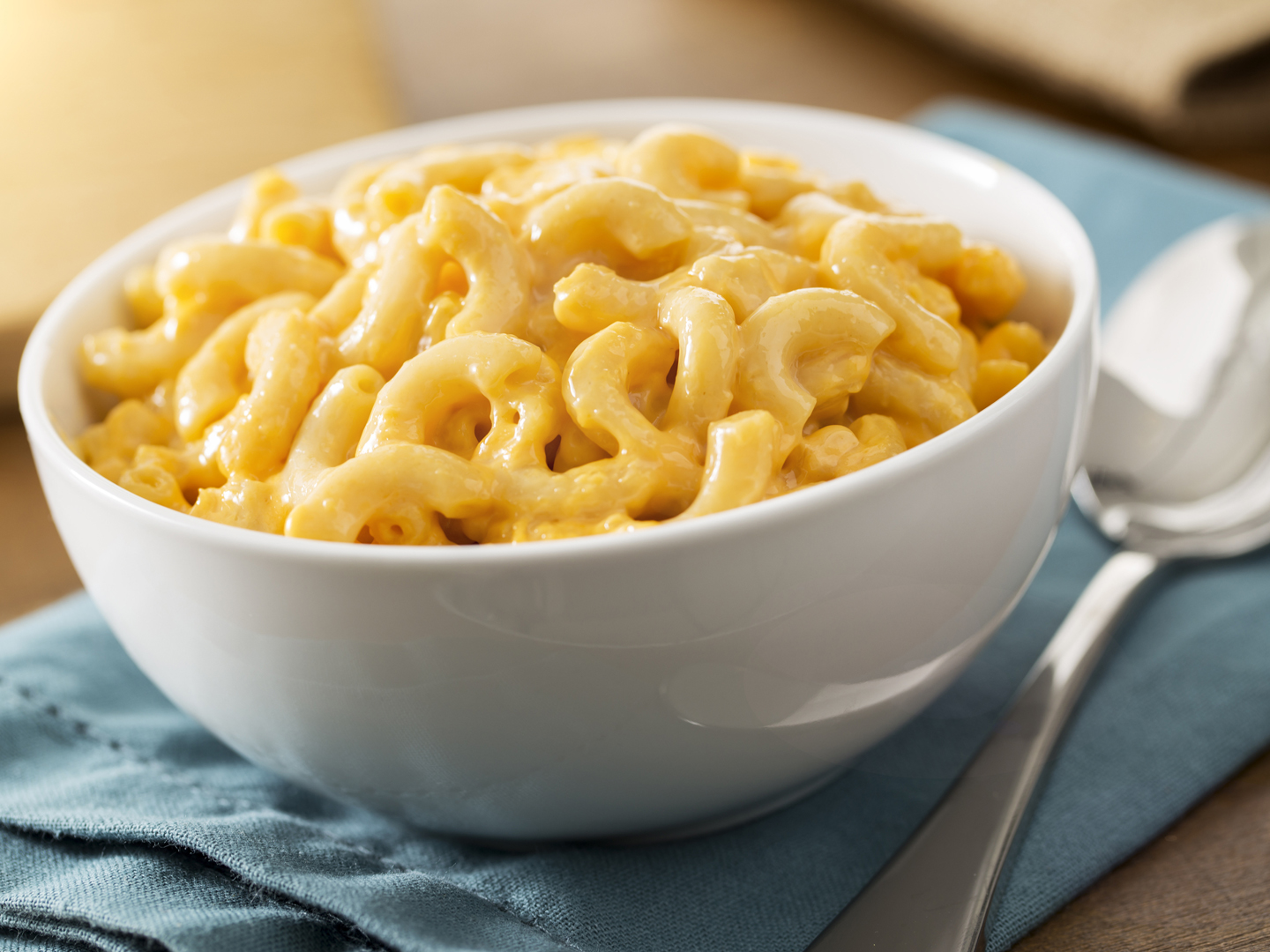 A white bowl of the best baked homemade macaroni and cheese on a table with a blue napkin.  The recipe is made with elbow pasta and cheddar cheese.  The shot is vertical and has selective focus.  Macaroni and cheese is a American comfort food or soul food.
