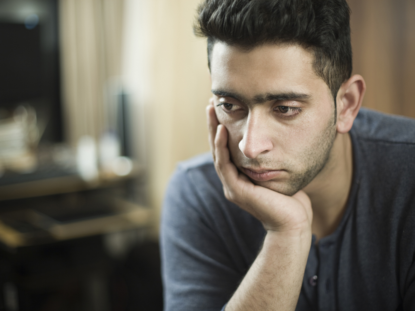Indoor low key image of a serene young man thinking by resting his head on hand and looking away with and blank expression. He is wearing a t-shirt. One person, waist up, horizontal composition with selective focus and copy space.