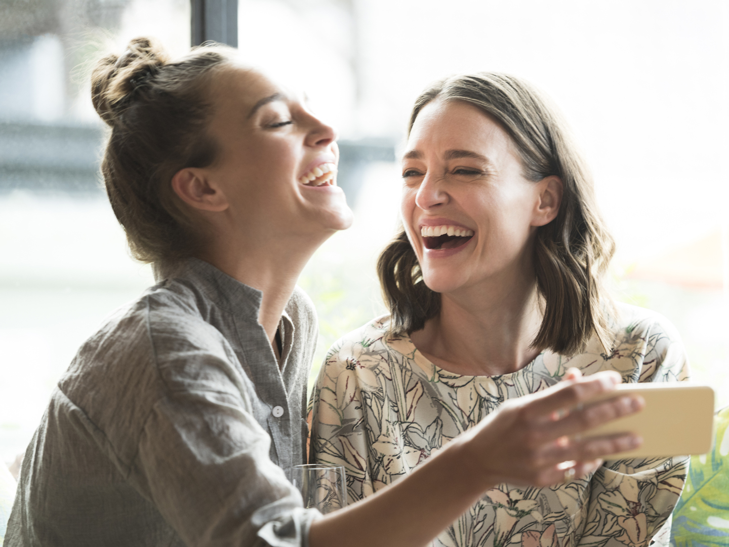 Two female friends with smartphone, smiling. Young woman holding phone, mid adult woman looking at her and laughing