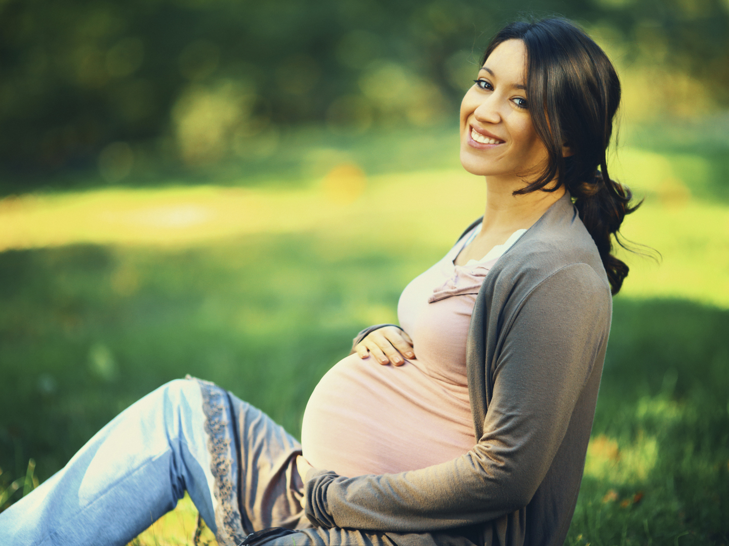 Closeup side view of smiling late 20&#039;s pregnant woman relaxing in local park on sunny summer afternoon. She&#039;s stroking her belly and looking at camera. Blurry trees and grass in background.
