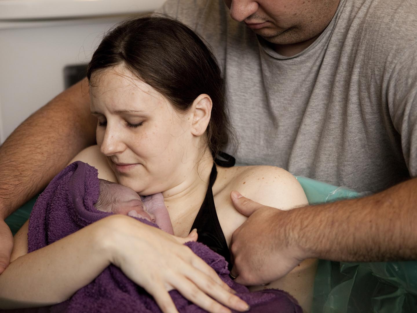 Color photo of a husband comforting his wife while she holds their newborn baby in the water of a birthing tub immediately after a water birth at home.
