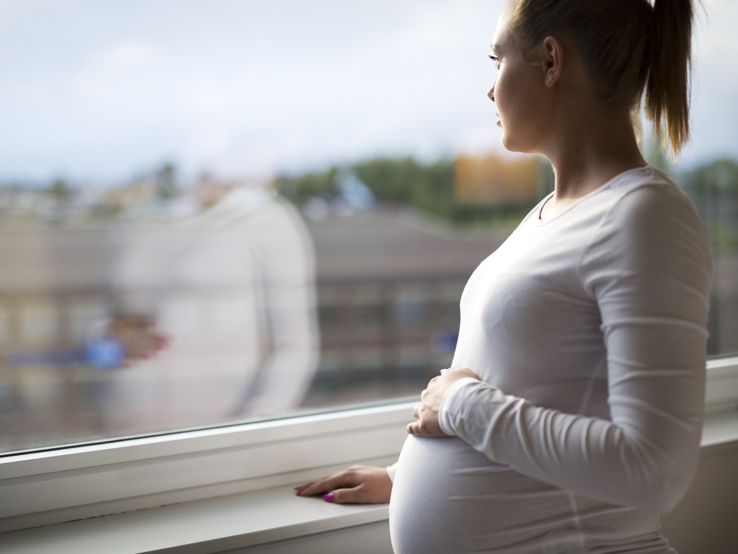 Thoughtful pregnant woman looks out the window home. Holding a hand on the tummy. Mental health and pregnancy.