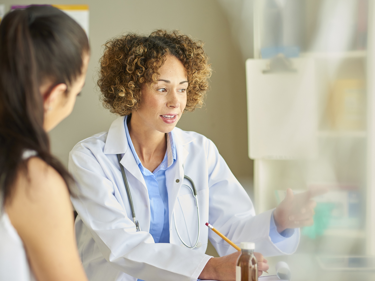 A female doctor sits at her desk and explains diagnosis to a female patient .  She is wearing a white coat and stethoscope and is referring to a computer screen whilst making notes.