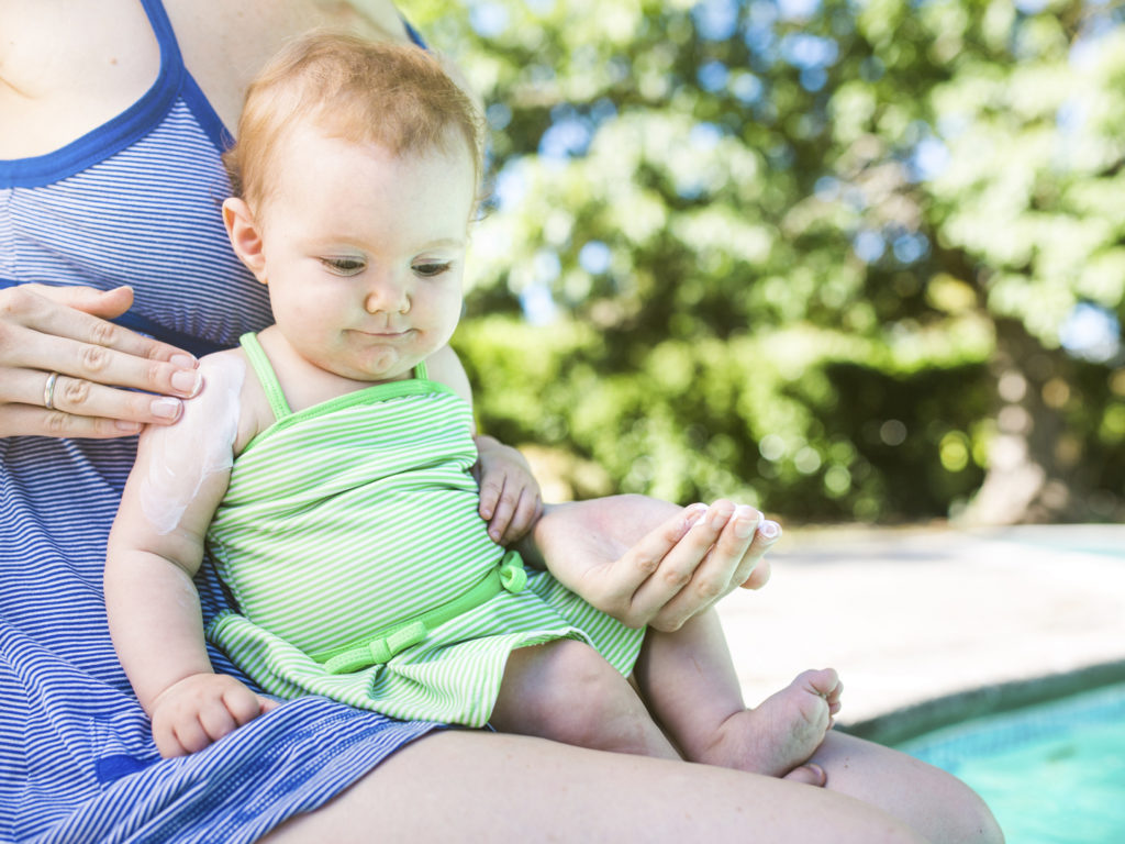 A close up of a mom and her smiling baby girl sit next to a pool, the mom applying sunblock to her daughters skin to protect from the suns harmful U.V. rays. The child smiles, patiently letting her put on the lotion. Horizontal image with copy space.