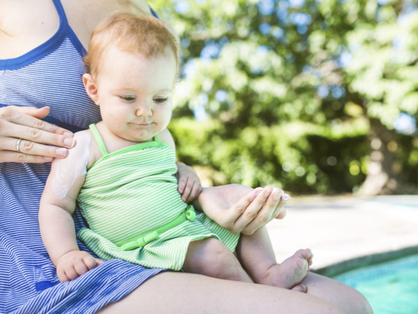 A close up of a mom and her smiling baby girl sit next to a pool, the mom applying sunblock to her daughters skin to protect from the suns harmful U.V. rays.  The child smiles, patiently letting her put on the lotion.  Horizontal image with copy space.