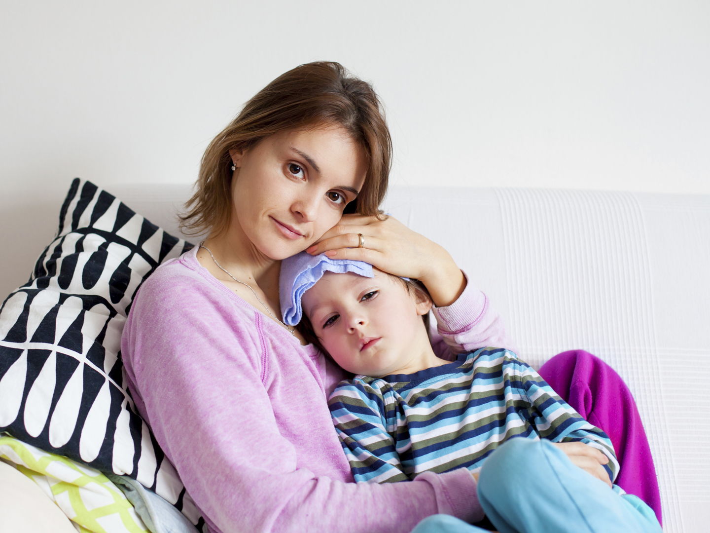 Young mother, holding her little sick boy, wet cloth on his forehead, trying to decrease his temperature