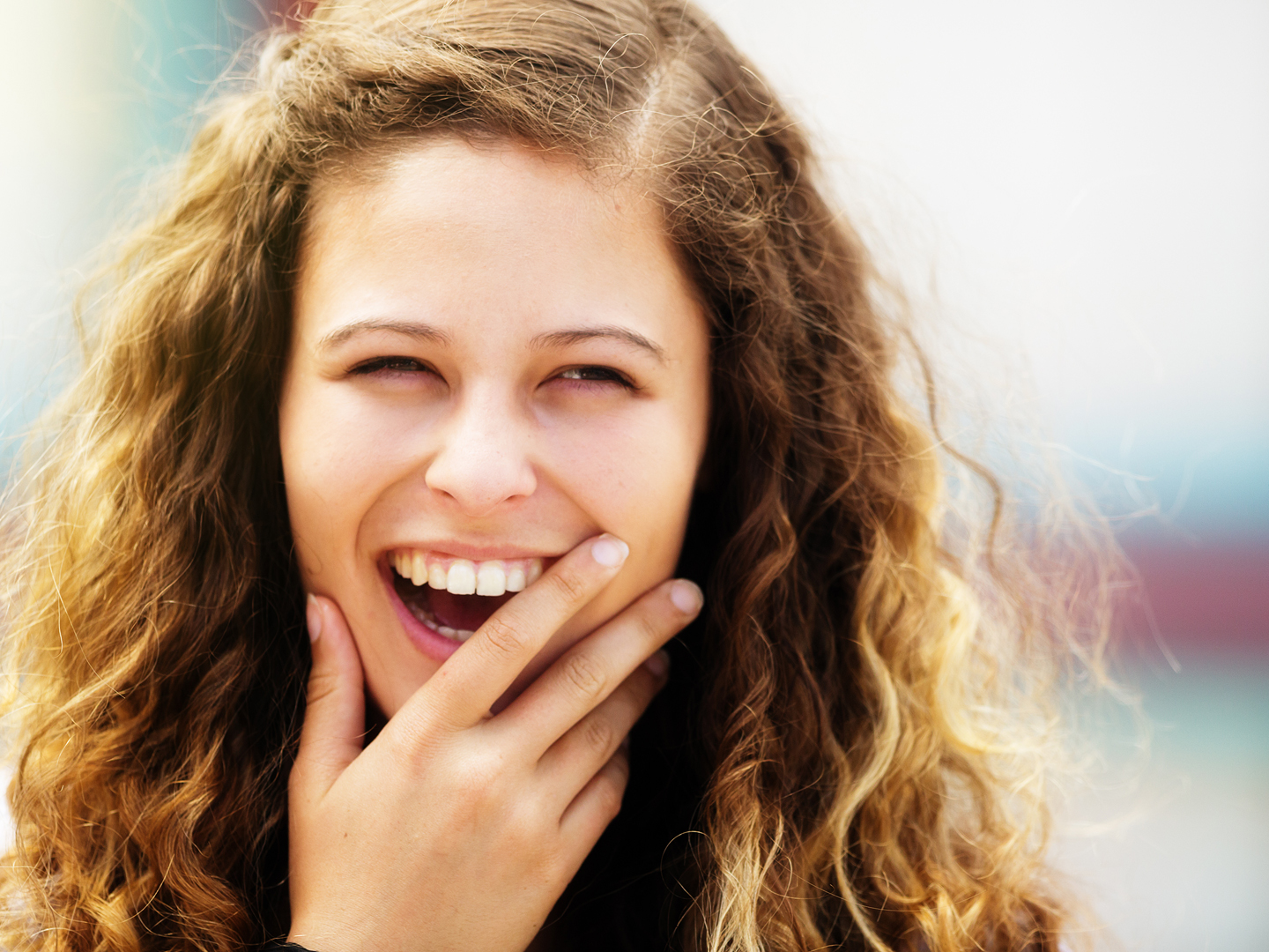 Head and shoulders portrait of a lovely young girl, with curly, honey-blonde hair and brown eyes, laughing happily in the sunshine with her hand partly over her mouth.