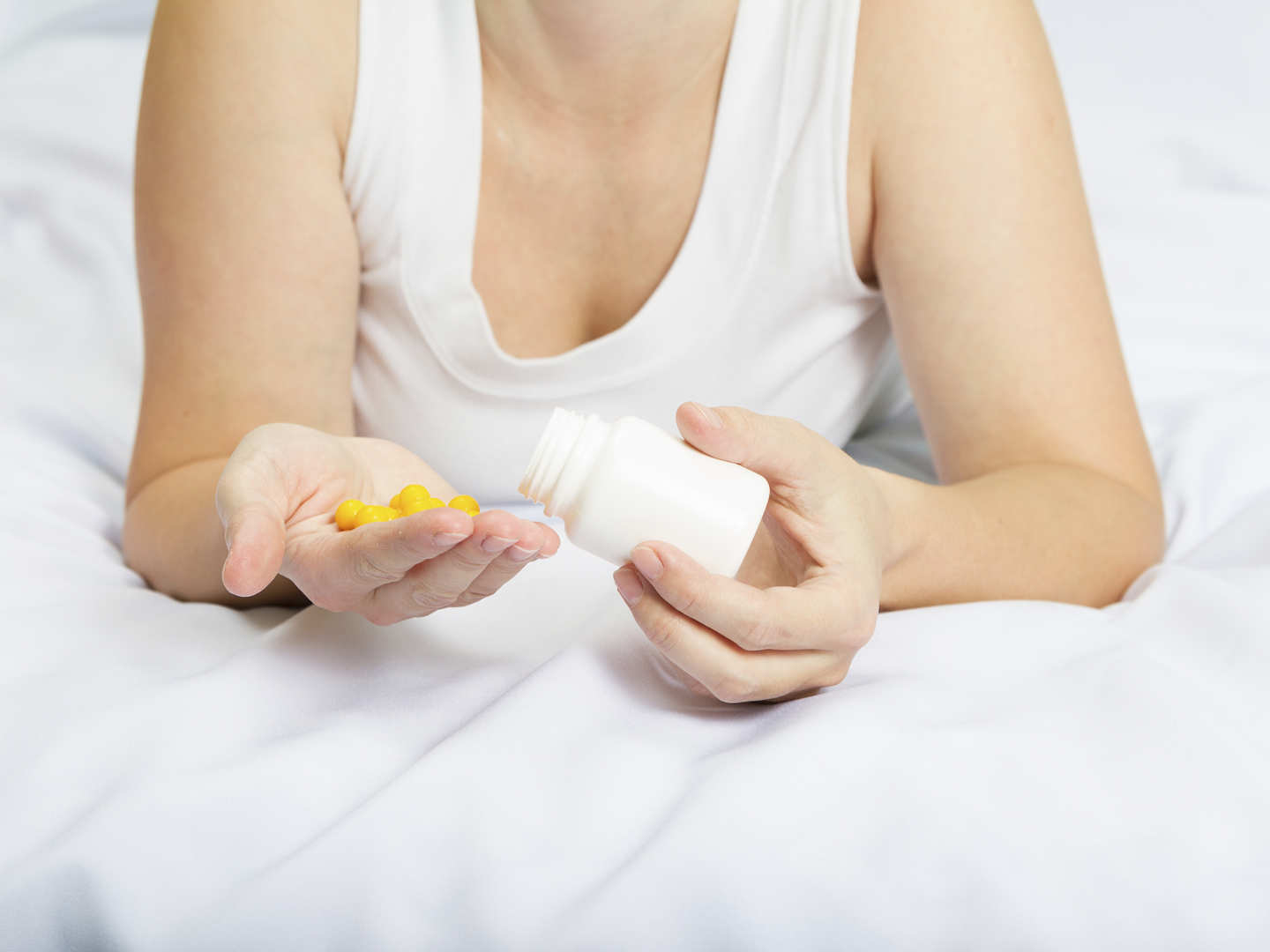 Young Caucasian woman in her pajamas in bed holding yellow pills from a white bottle.