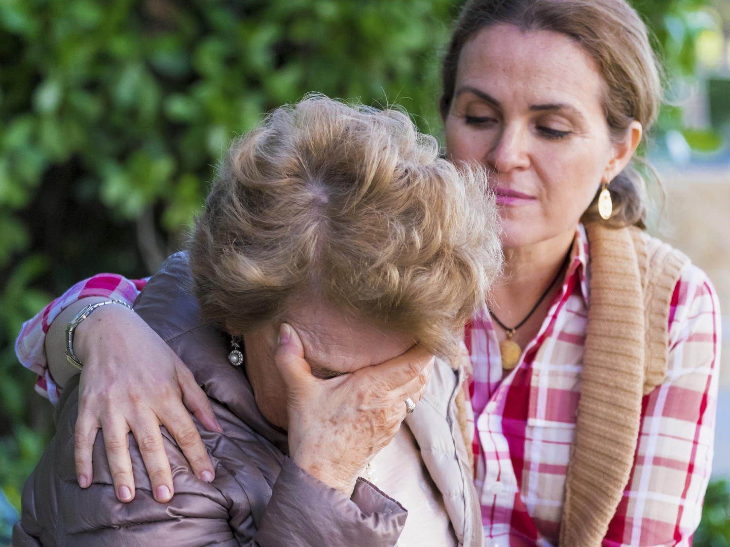 Senior woman with her hands on her head looking down sitting in a park bench with her daughter consoling her