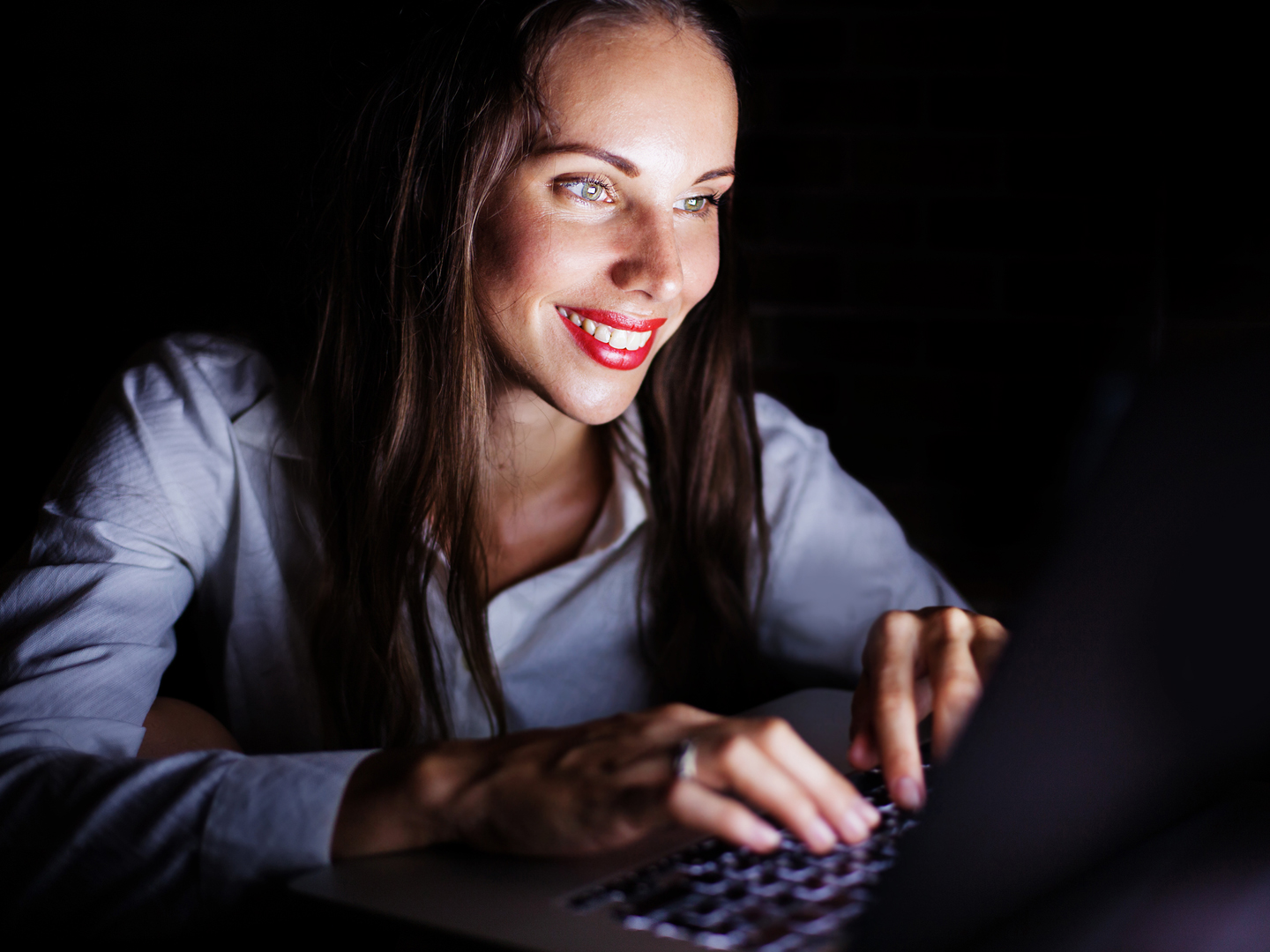 Beautiful caucasian women in white shirt sitting in dakness and typing on laptop with smile
