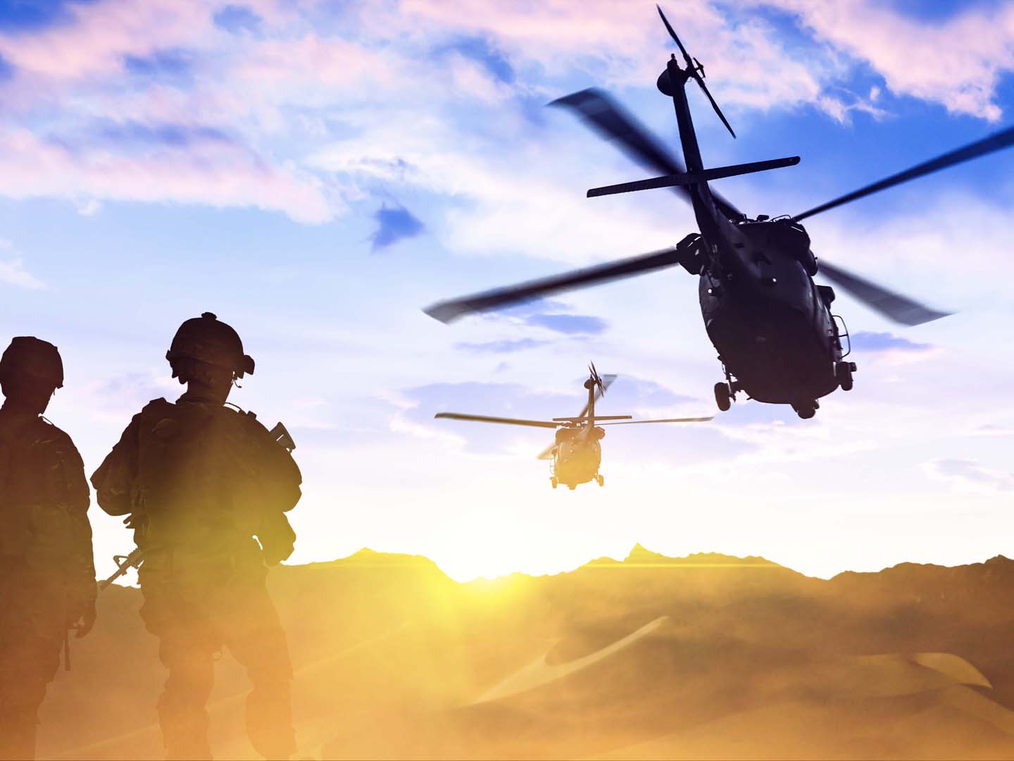 A military helicopter flies over troops on the ground at sunset.