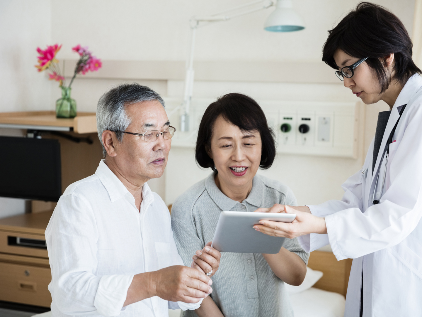 Female doctor showing digital tablet to senior couple sitting on bed in ward