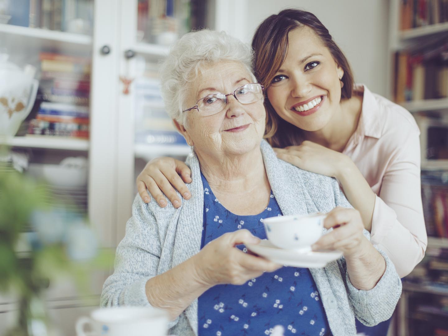 Social worker is visiting a senior woman in her own apartment. They are celebrating the elderly lady&#039;s birthday. The kind nurse is hugging the senior woman over her shoulder.