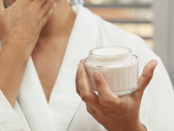 Close up portrait of mature woman applying facial cream. She is wearing white bathrobe. Selective focus. Face is not visible.