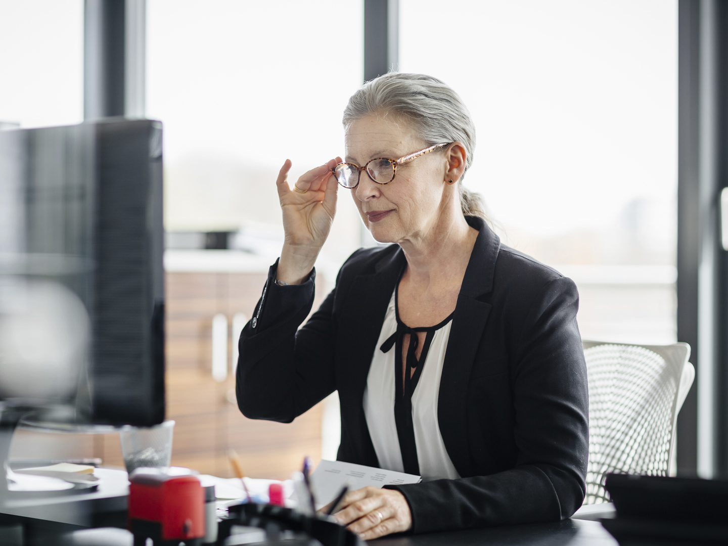 Portrait of a senior businesswoman sitting on her desk in a modern office and. She&#039;s working on a computer and wearing glasses and has silver hair.