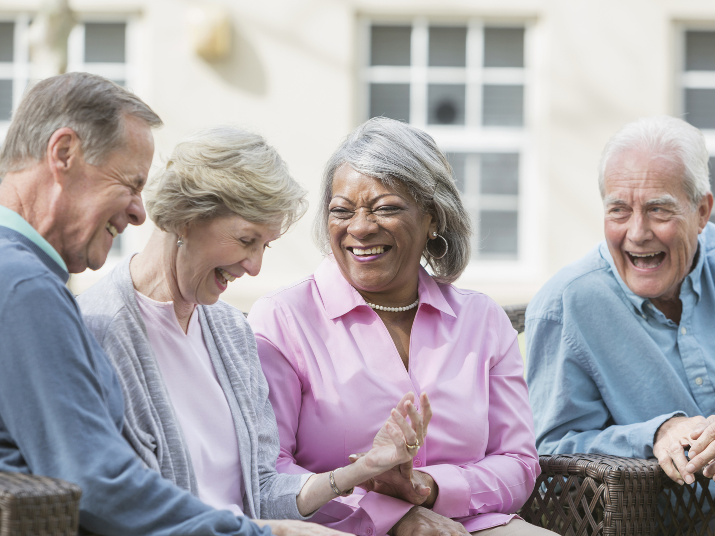 A group of four multi-ethnic seniors sitting together on patio furniture outdoors, talking and laughing. They are in retirement, relaxed and enjoying spending time with friends.