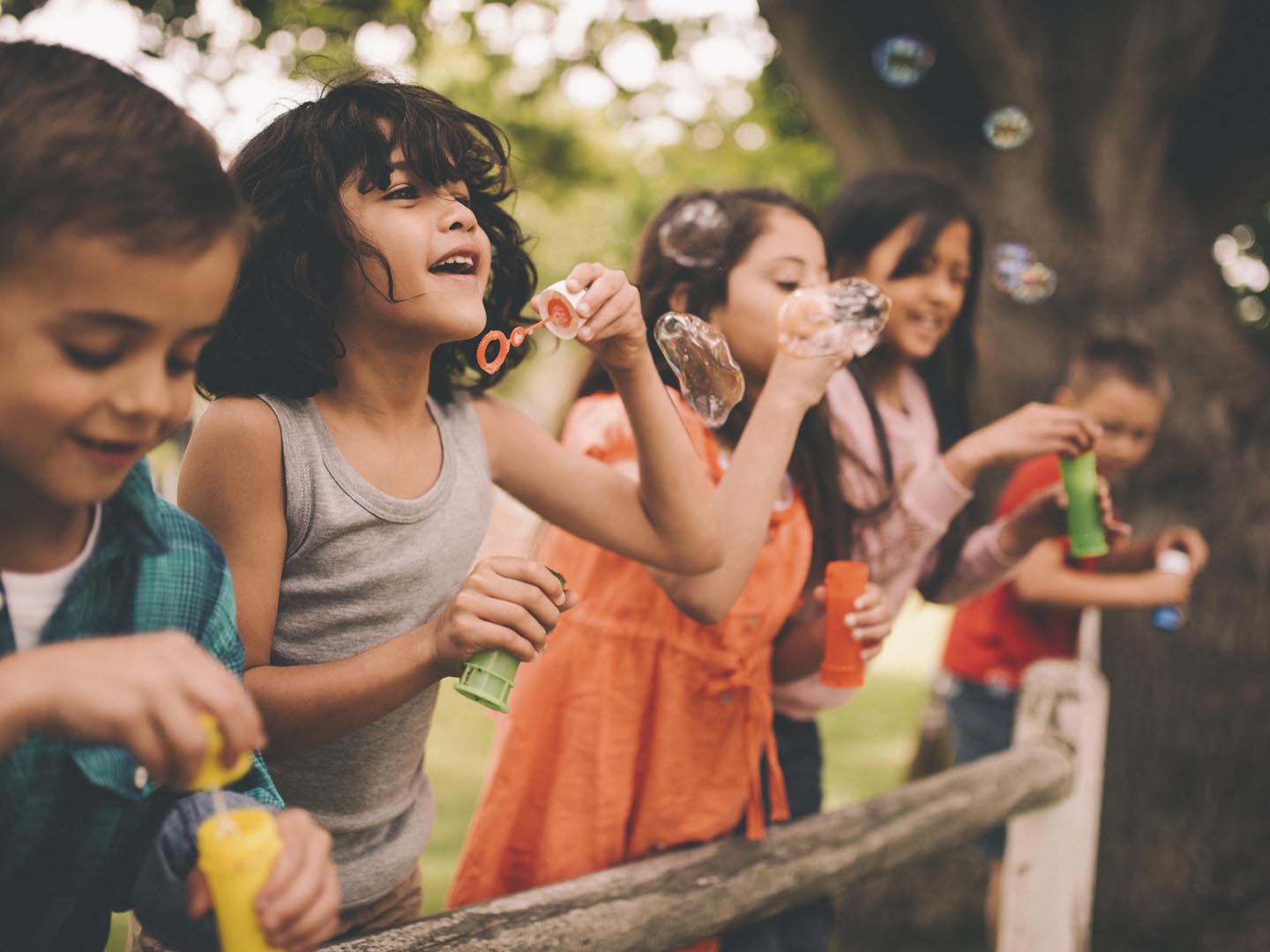 Long haired hispanic boy laughing and having fun with his friends standing on a wooden fence in a summer park blowing bubbles, with a vintage develop
