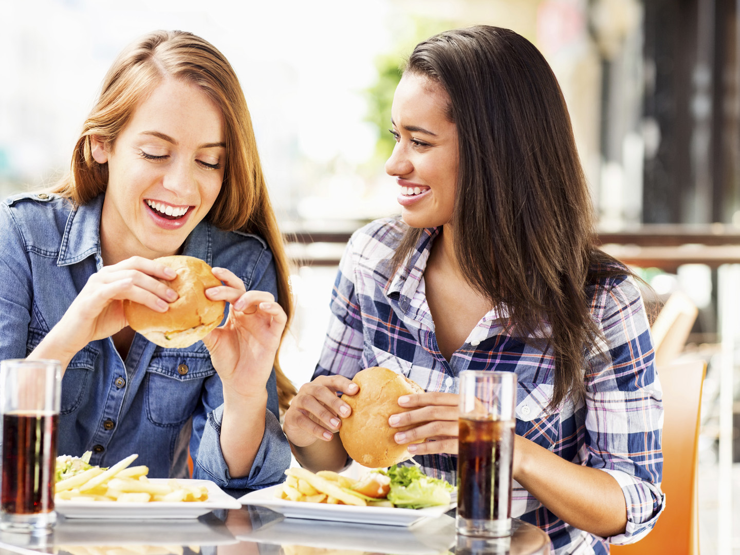 Two teenage girls eating hamburgers at a fast food restaurant.  The girl on the left has long reddish-brown hair and is wearing a denim jacket.  The girl on the right has long dark brown hair and is wearing a blue-and-white plaid shirt.  Each girl is holding a hamburger over a plate of French fries with lettuce on the side.  The two plates are square and white, and each one is sitting next to a soft drink in a tall, clear glass.  The girls are sitting side-by-side at a small, round table.
