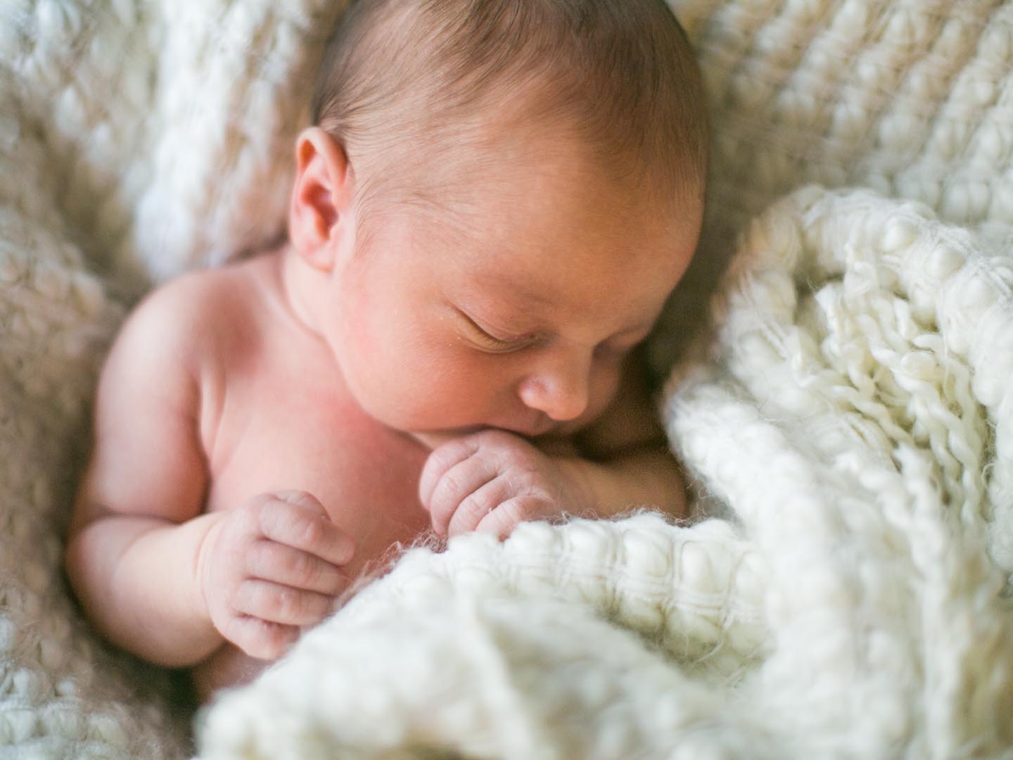 A brand new one week old baby boy sleeps wrapped in a white fuzzy blanket. He is peaceful and his hand rests near his mouth.