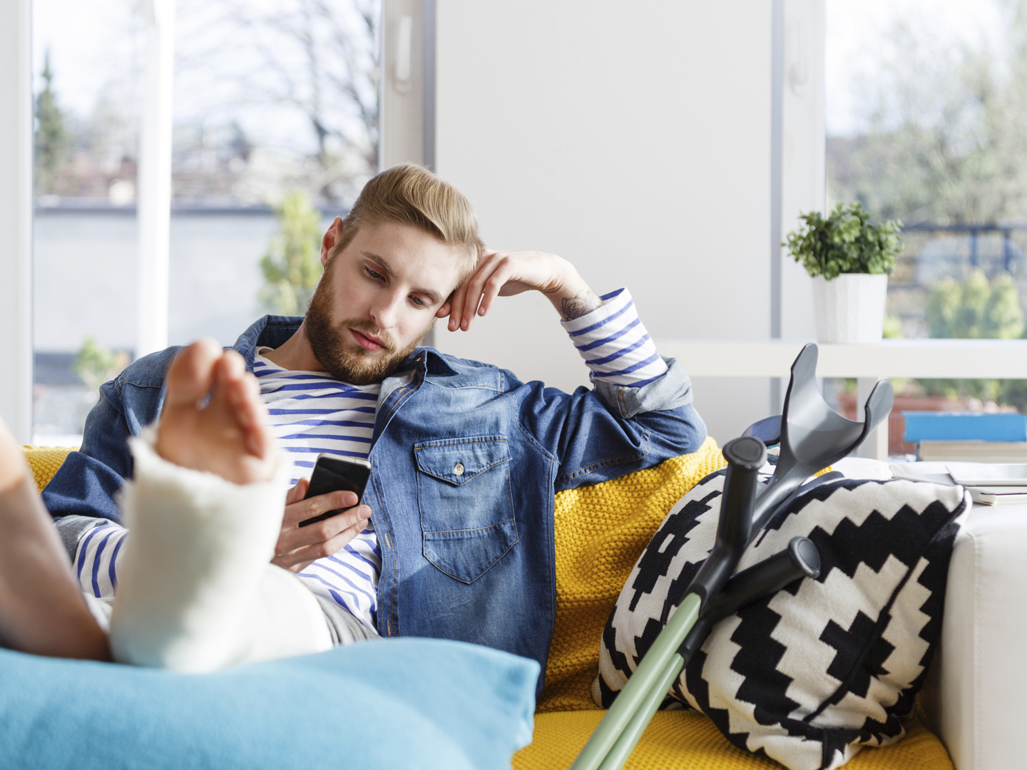 Young man with broken leg in plaster cast lying down on sofa at home and using a smart phone.