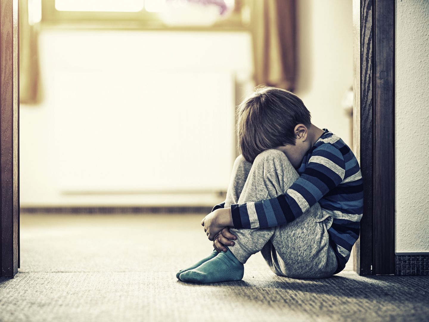 Depressed sad child sitting on the floor, in the door. The little boy is hiding his head between legs.
